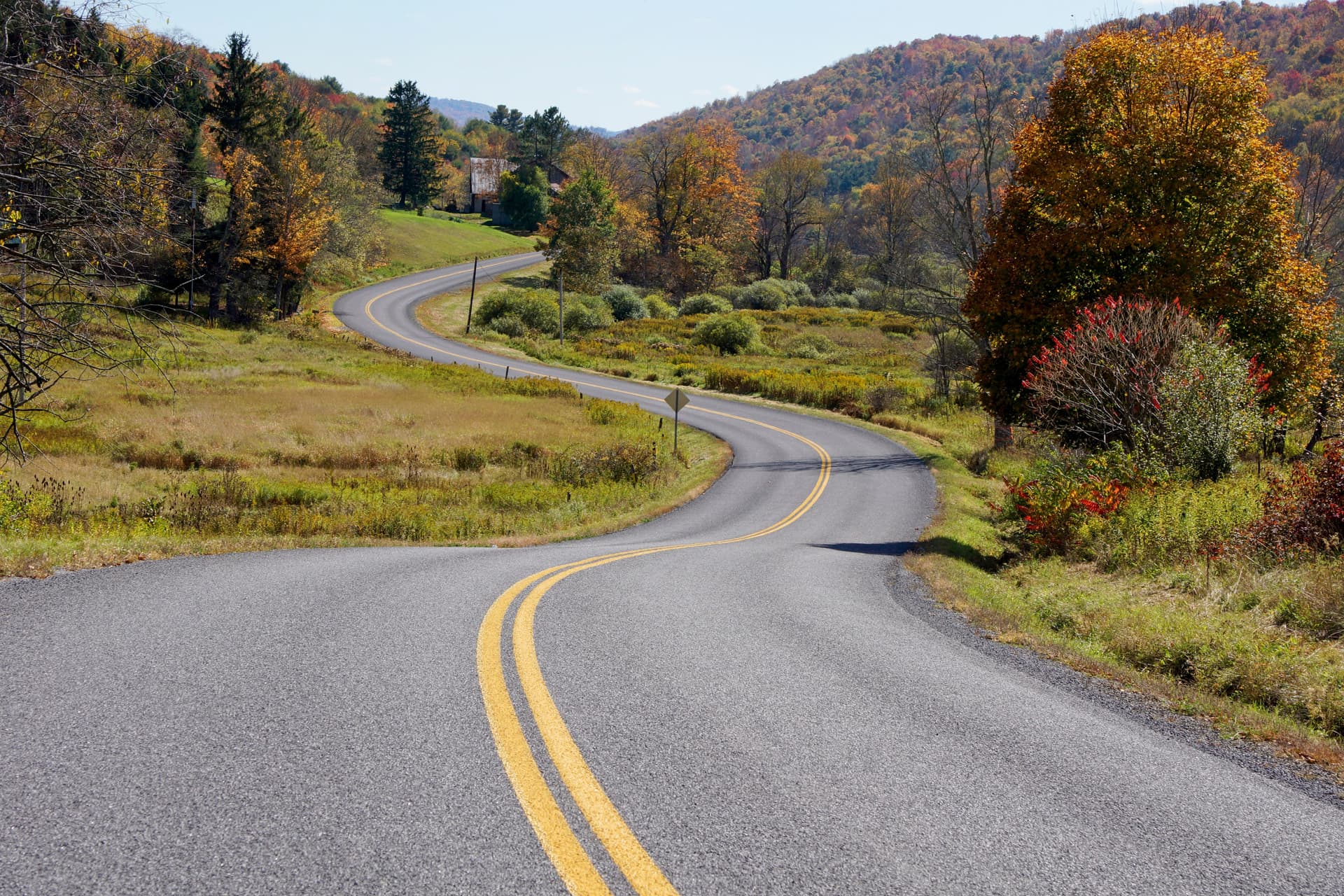 A winding road through the Hudson Valley in autumn, with the Catskill Mountains visible in the distance and mature trees lining the roadside.