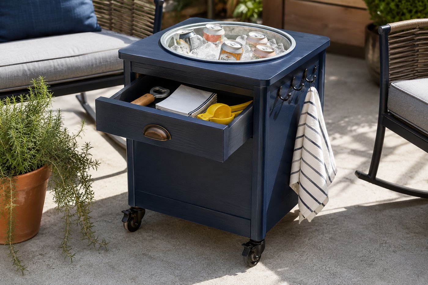 Thrift store nightstand painted in deep navy blue on a sunny patio, with a galvanized metal bucket dropped into a cut hole in the top surface filled with ice and drinks, the drawer slightly open showing a bottle opener and napkins, and casters visible at the base
