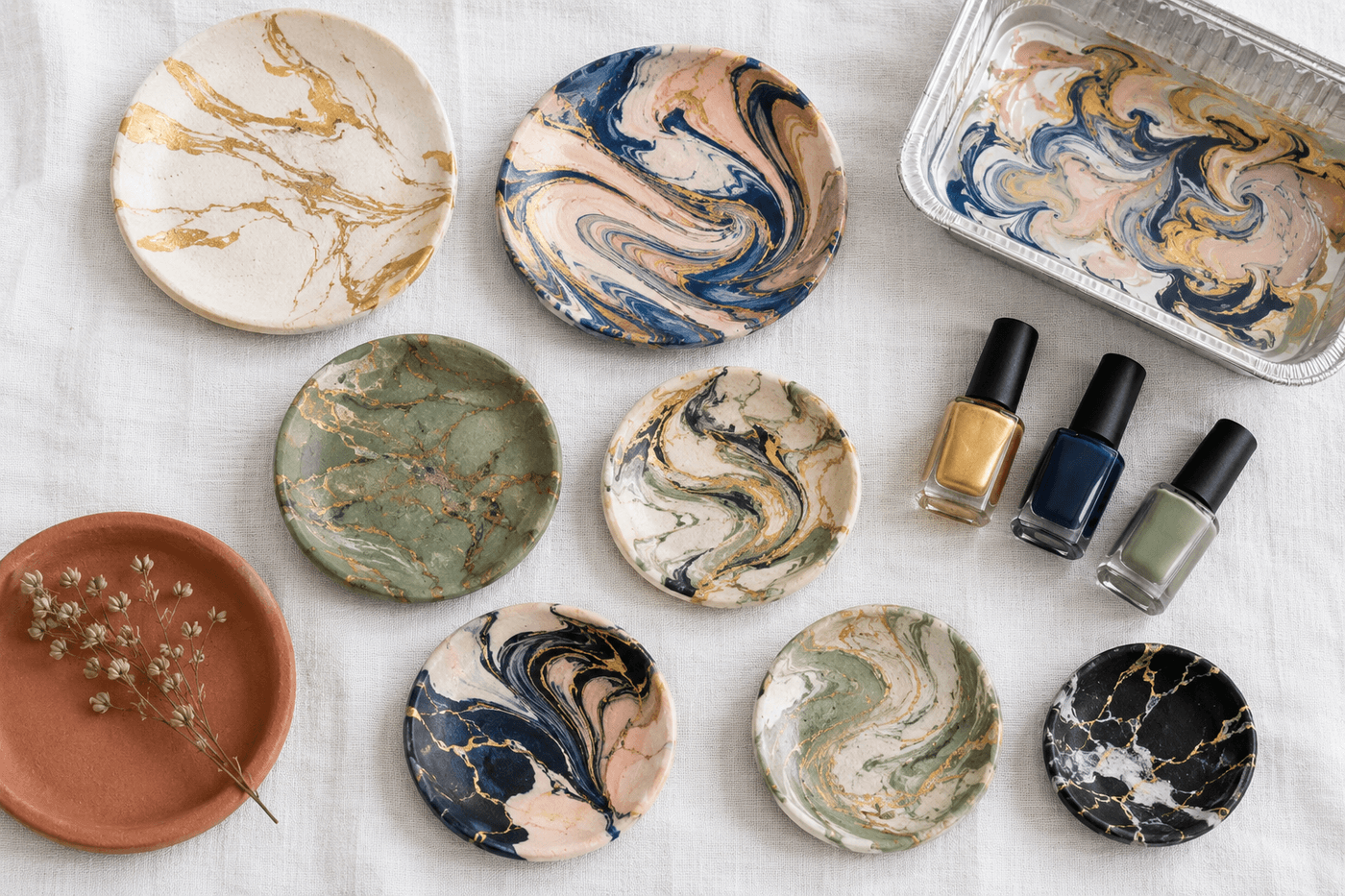 Eight small air-dry clay trinket dishes in varying sizes displayed on a white linen surface, each showing a unique marble pattern in gold, navy, sage green, and blush tones transferred with nail polish marbling technique
