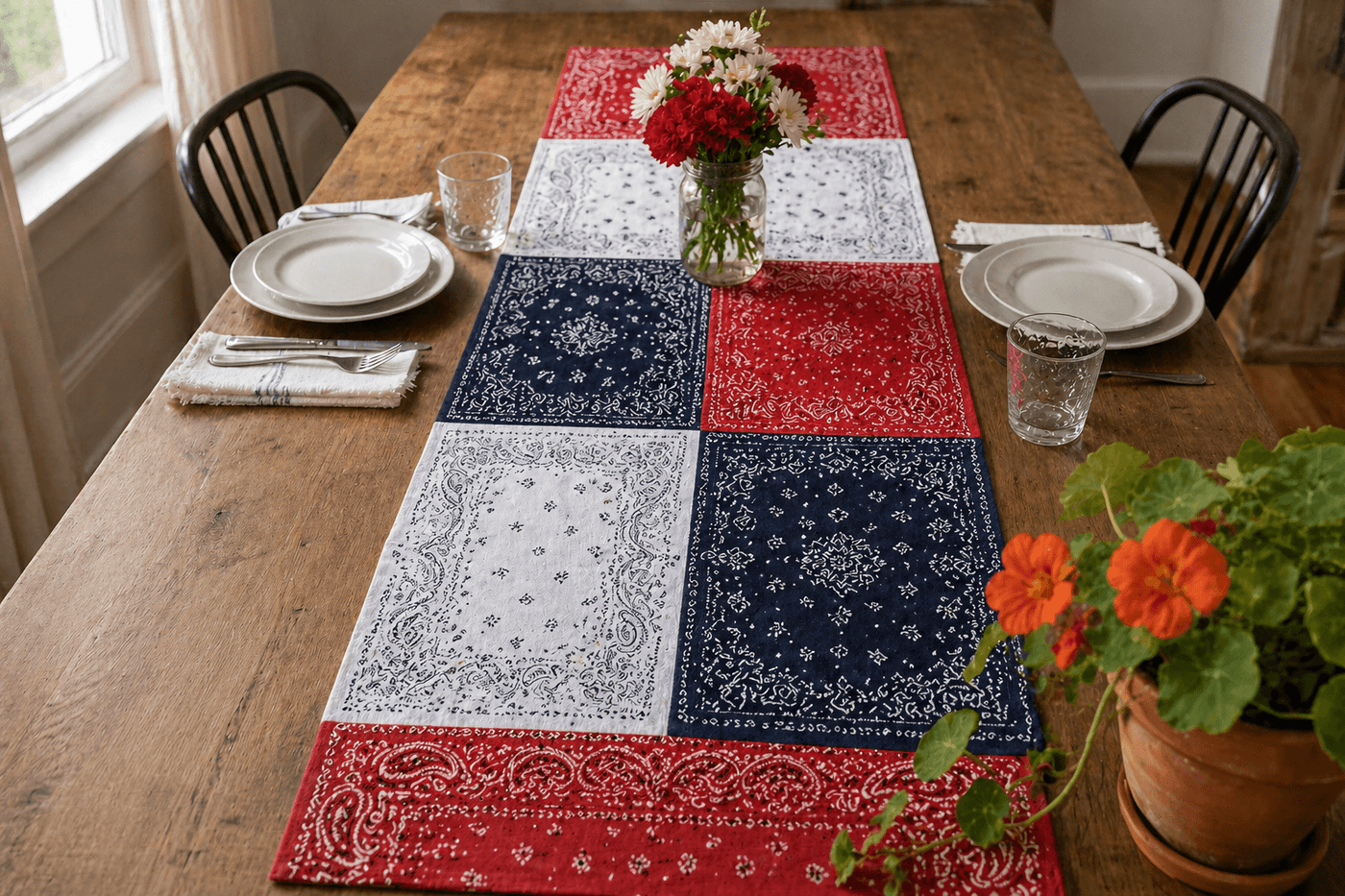 A quilted bandana table runner in red, white, and blue paisley bandanas sewn together in two columns running down a wooden farmhouse table, styled with white ceramic dishes and small flag accents