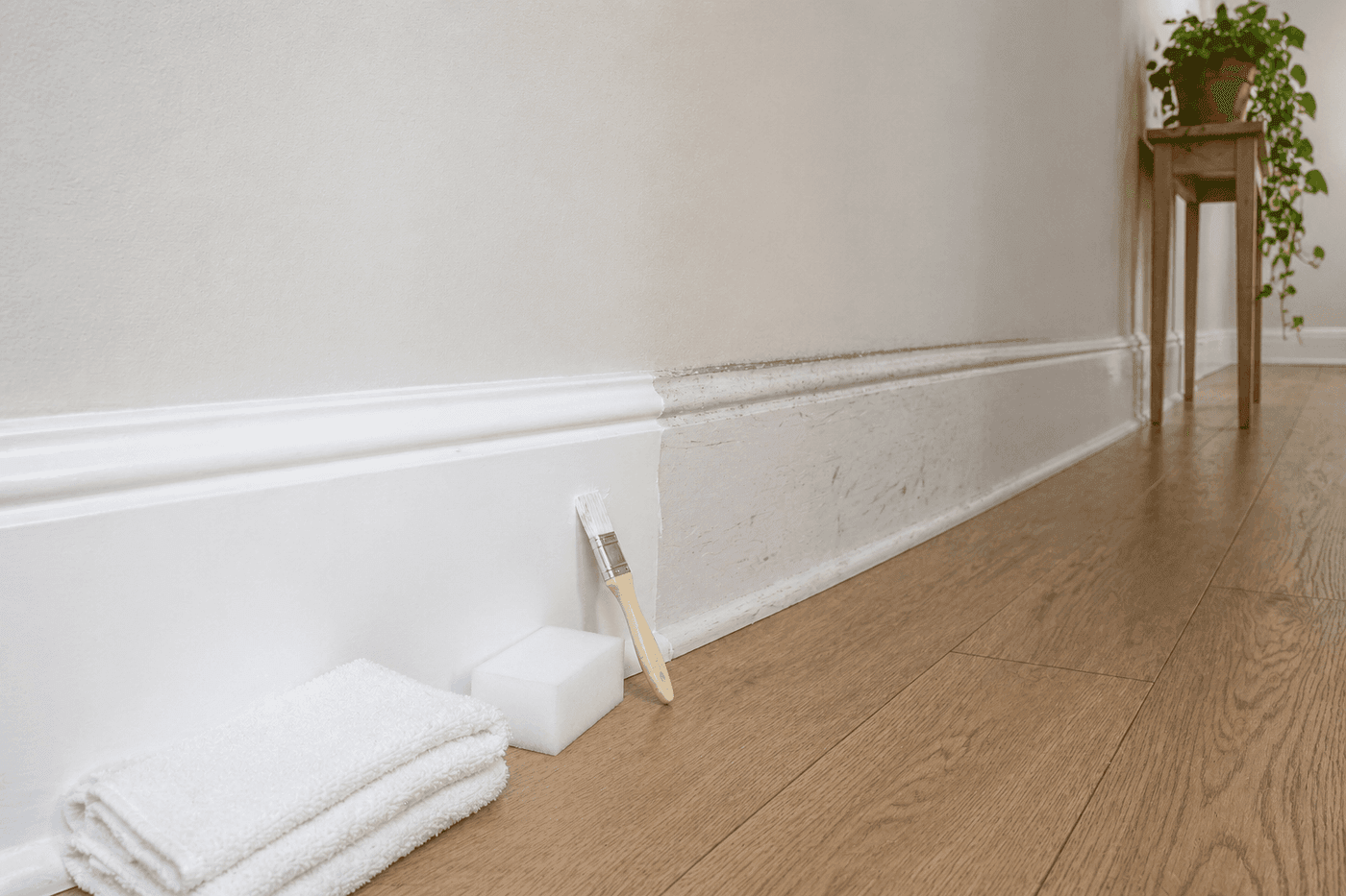 Clean white baseboards in a bright hallway after wiping, showing the contrast between a freshly cleaned section and a dusty uncleaned section beside it, with a microfiber cloth and small trim paint brush on the floor nearby